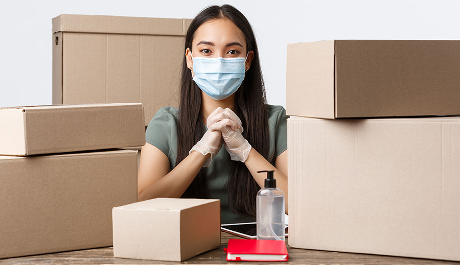 A woman surrounded by empty boxes wearing a mask, gloves, and with a hand sanitizer in front of her. 